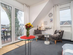 Una habitación moderna con una mesa de vidrio, dos sillas con diseño, un sofá, una mesa baja de vidrio, un jarrón con flores, un reloj y vistas al exterior.
