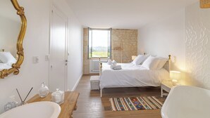 A bedroom with a large bed, a white bathtub, a wooden bench, a mirror, and a stone wall.
