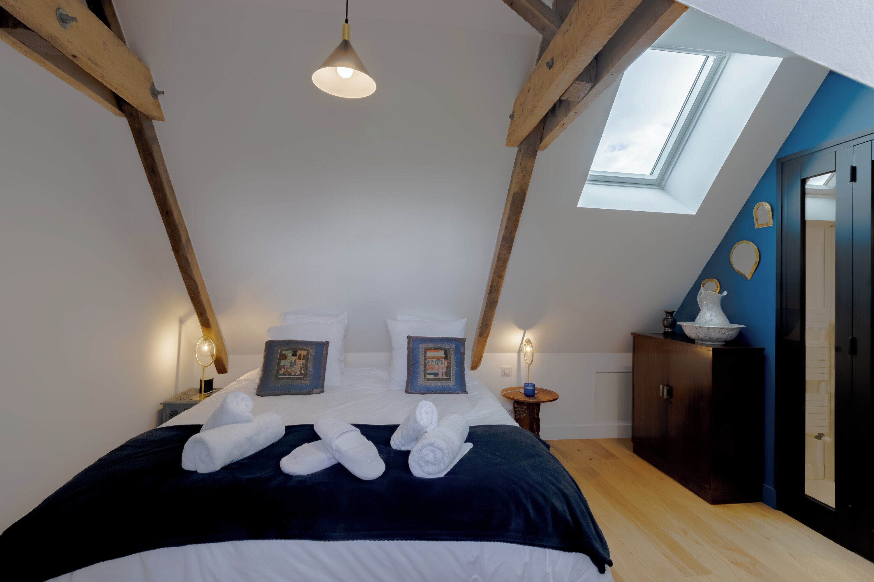 Un dormitorio con tragaluz, una cama con toallas ordenadas, una mesita de noche con lámpara y un armario de madera.