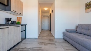 Eine moderne Küche mit Holzschränken, einer Kaffeemaschine und einem Geschirrspüler. Ein Gang führt zu einem Wohnbereich mit einem grauen Sofa.