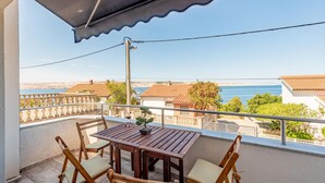 Ein Balkon mit Holztisch und Stühlen, der Blick auf die Küste mit Häusern und dem Meer freigibt.