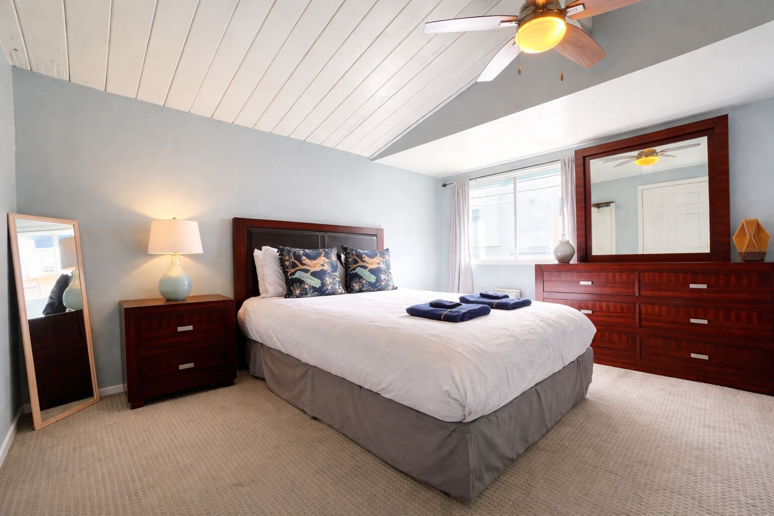 Une chambre à coucher comprenant un lit, des tables de chevet, une commode, un ventilateur de plafond et un grand miroir.