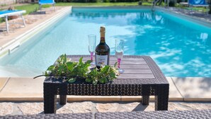 A poolside table with a bottle and glasses, surrounded by a pool and greenery.