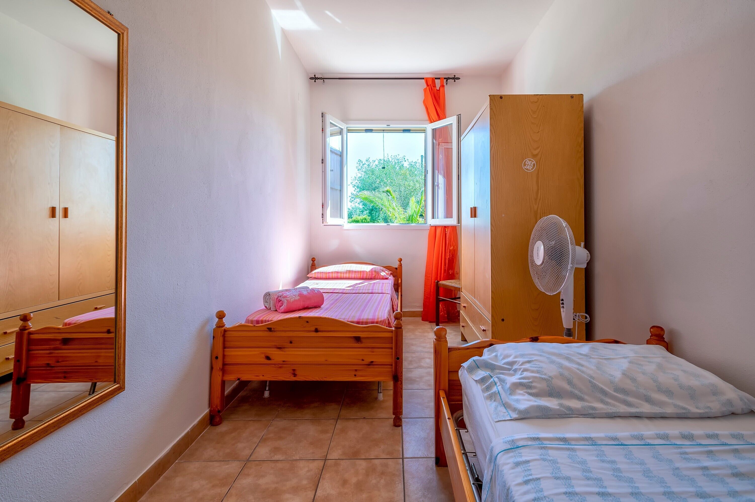 Une chambre à coucher avec un lit en bois, une table de chevet, un miroir, une fenêtre avec des rideaux et un ventilateur.