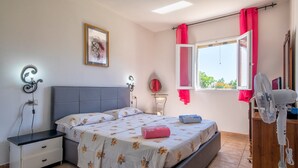 A bedroom with a bed, bedside table, nightstand, window with curtains, and a fan.
