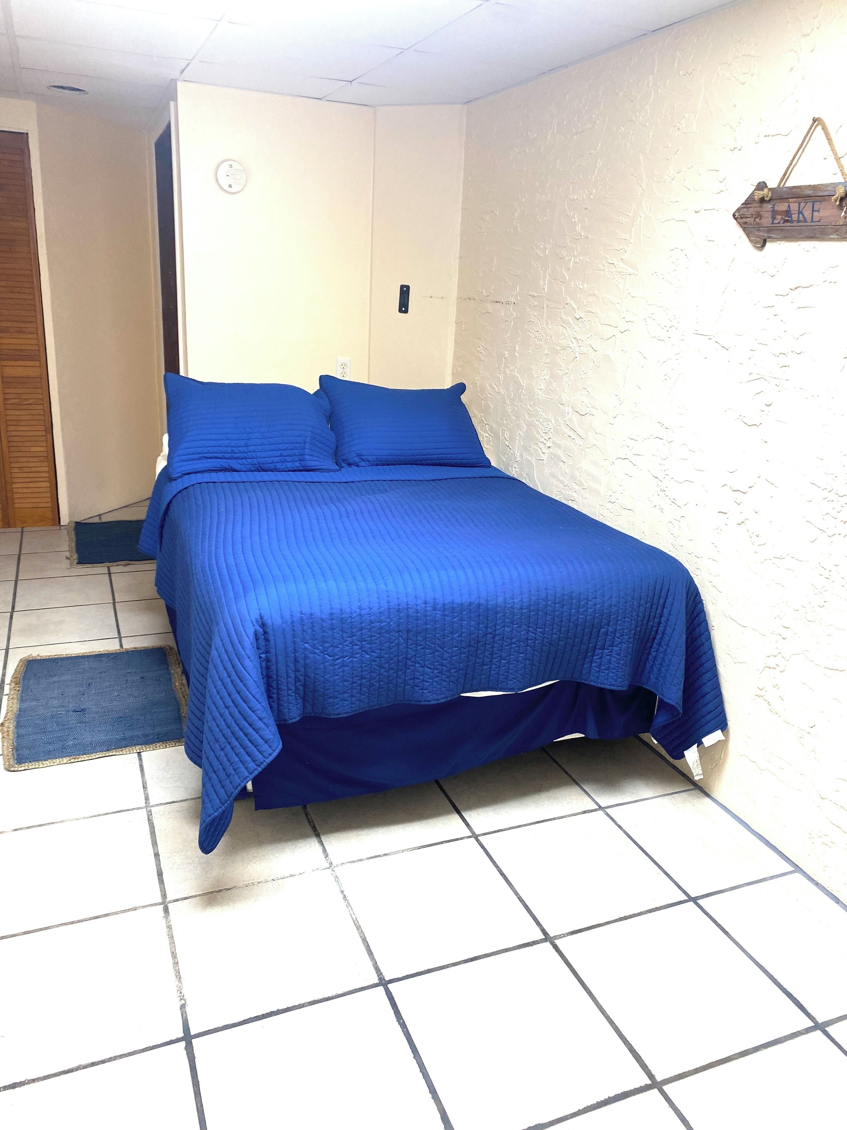 A room with a bed covered in blue bedding, a wooden door, a wall clock, and a sign that reads "LAKE".