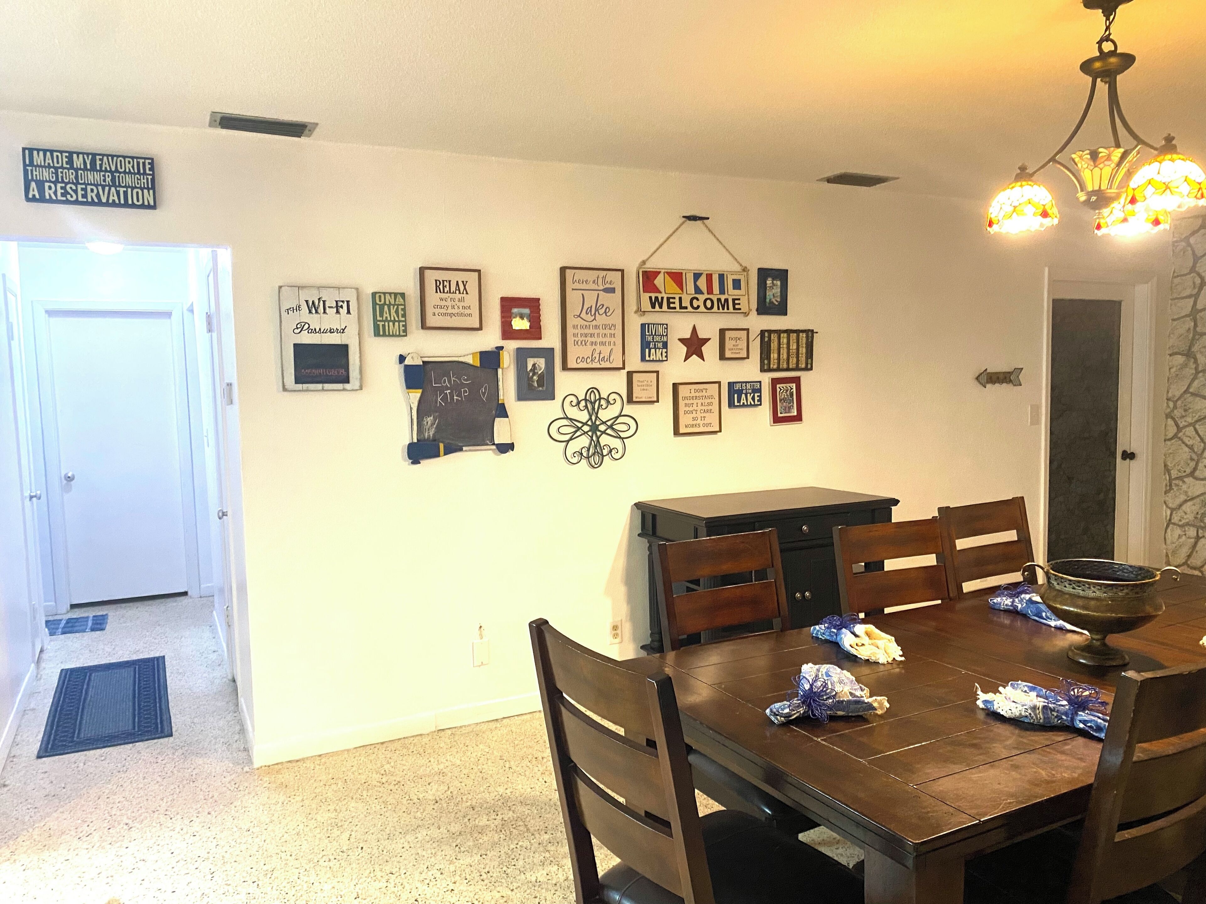Un comedor con mesa de madera, sillas y una araña. Hay cuadros y carteles en la pared, incluyendo un cartel de 'Welcome' y uno que dice 'Relax'.