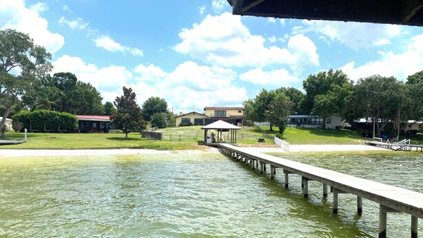 A calm lake with a wooden pier extending into it, surrounded by greenery and a few buildings in the background. - Lake Placid Vacation Destination for the whole family! (Lake Placid)