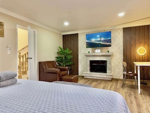 Une chambre d’hôtel comprenant un lit, un fauteuil en cuir marron, un téléviseur fixé au-dessus d’une cheminée, et un bureau avec une chaise.