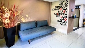 A modern hotel lobby with a grey sofa, a decorative plant, and a wall with multilingual greetings. - S06 PREMIUM SUITE Full Service WiFi Kitc (Atizapan de Zaragoza)