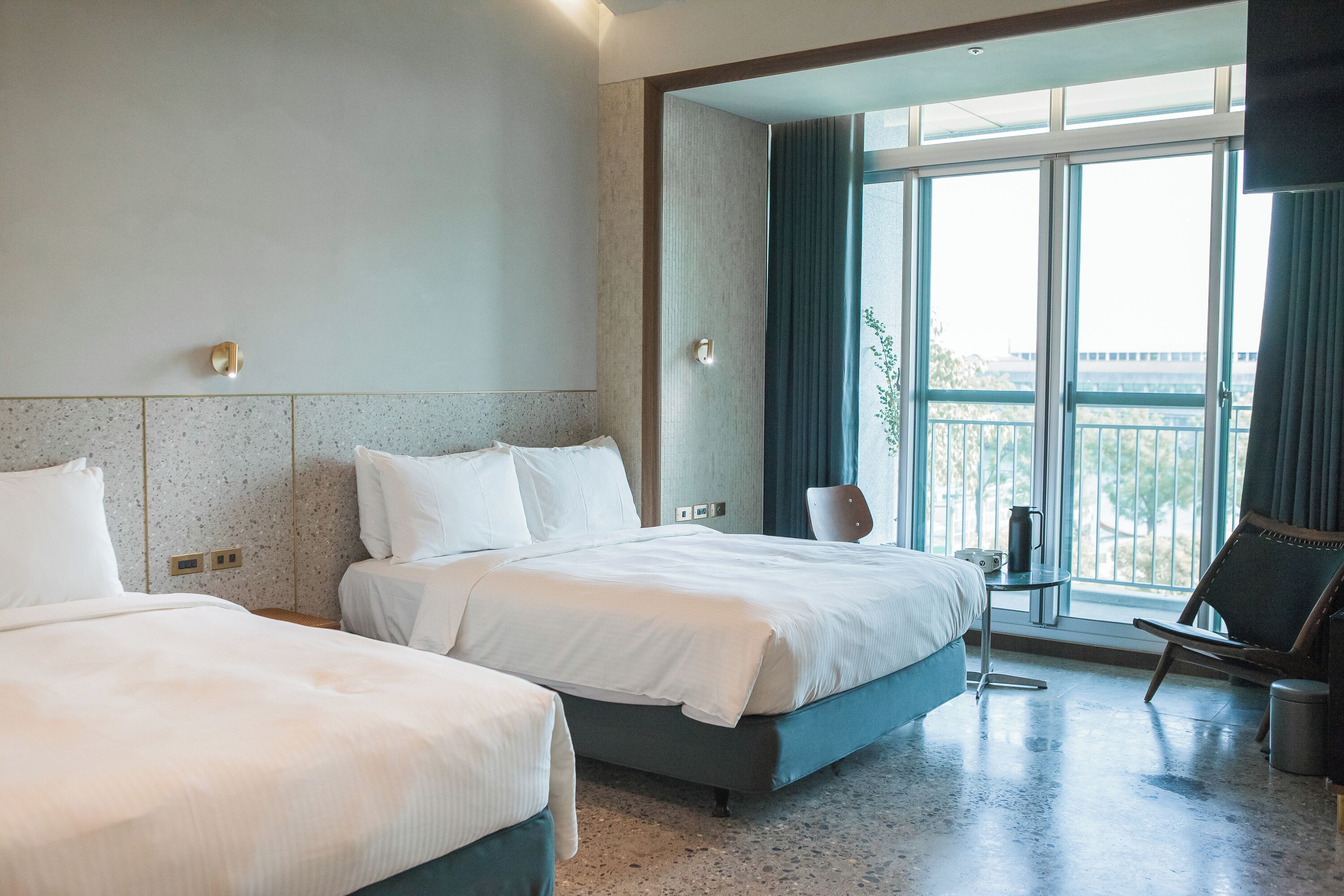 A hotel room with two beds, a small table, a chair, and a large window with a view.