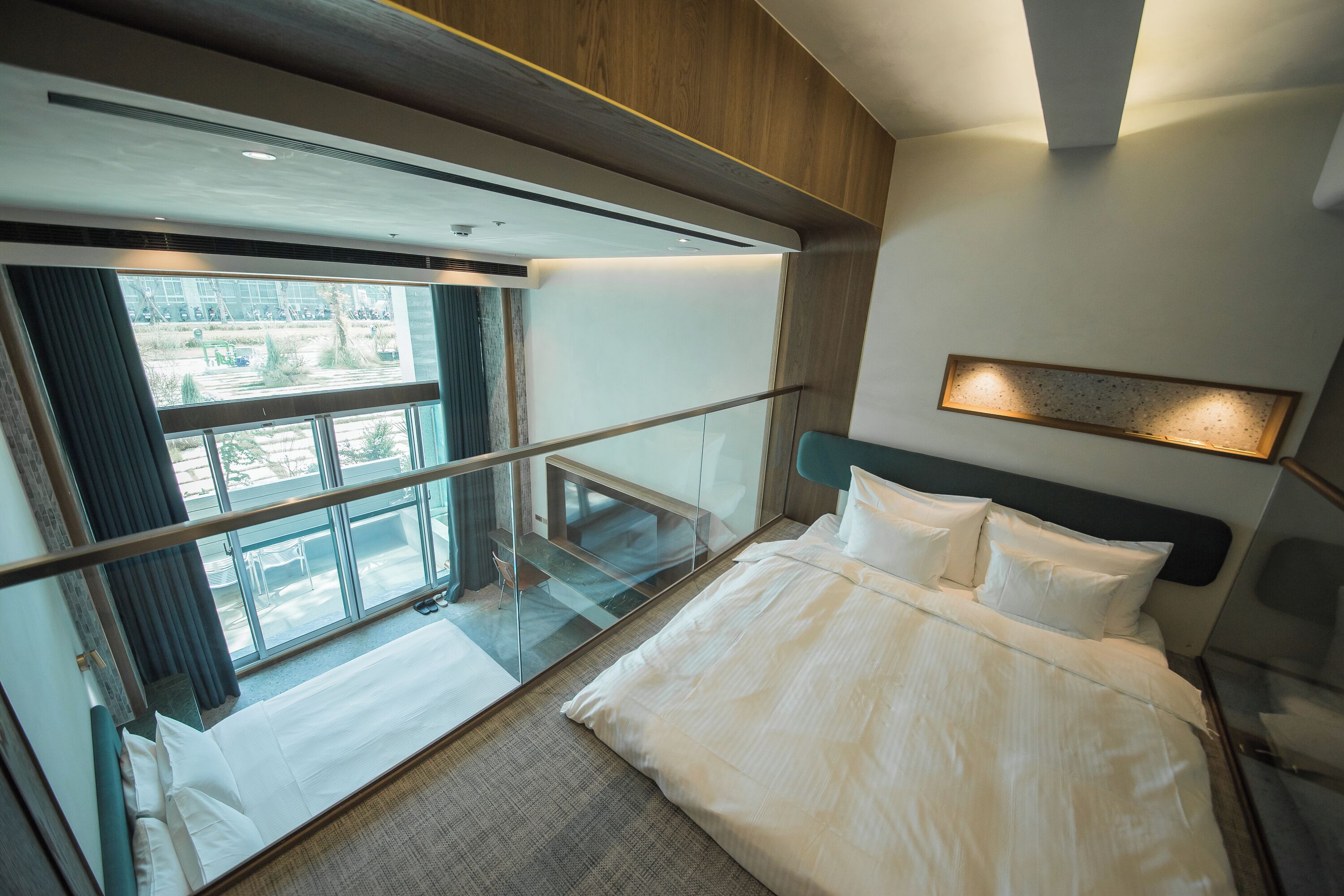 A modern hotel room with a large bed, a glass balcony, and a view of the city.