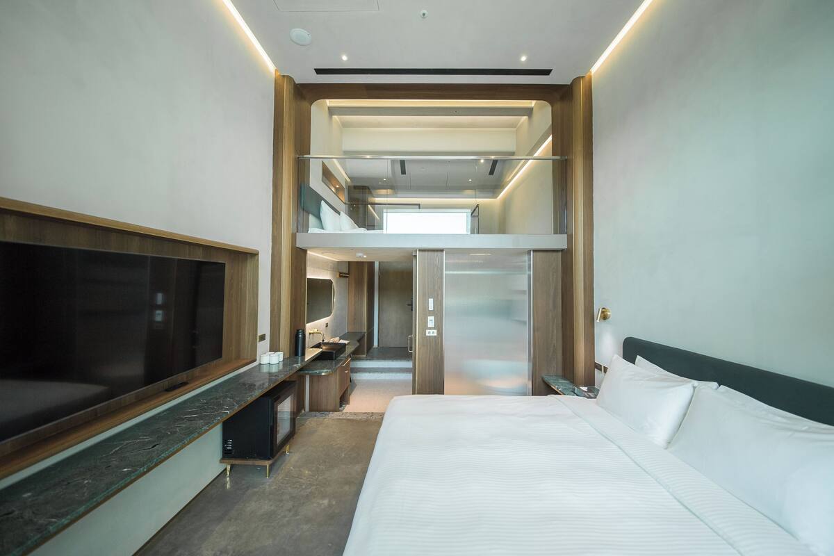 A modern hotel room with a large bed, a flat-screen TV, a desk, and a glass-enclosed balcony.