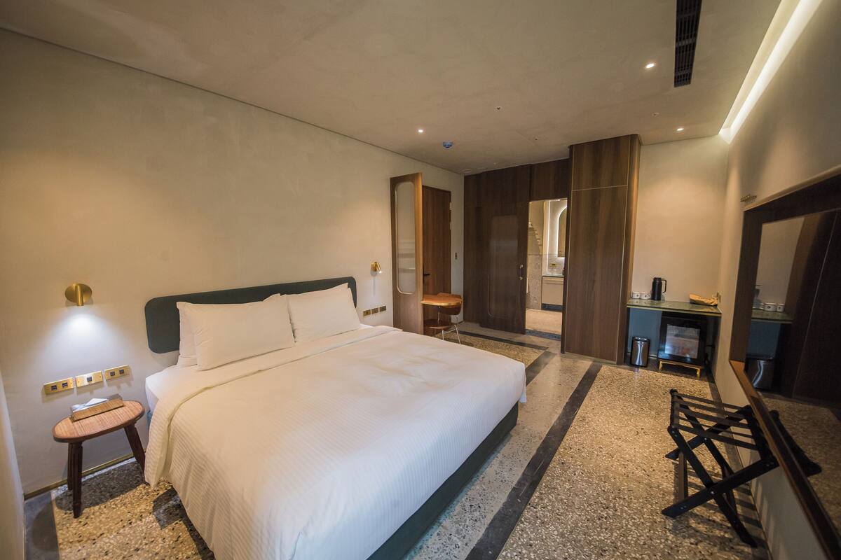 A hotel room with a large bed, a small stool, a desk, and a mirror.