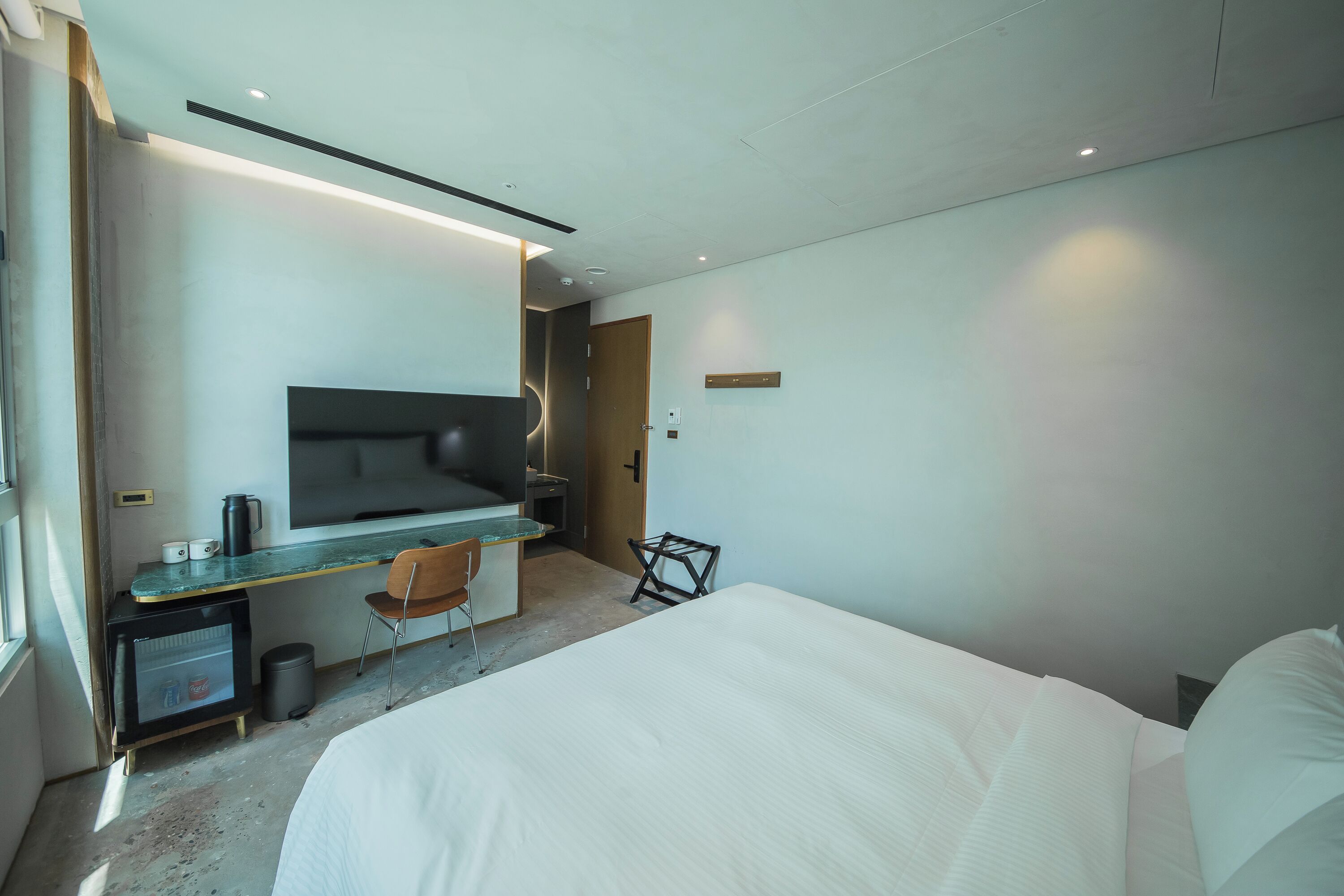 A modern hotel room with a bed, a desk with a chair, a small refrigerator, and a television.