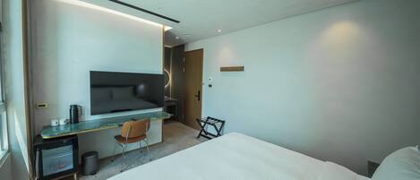 A modern hotel room with a bed, a desk with a chair, a small refrigerator, and a television.