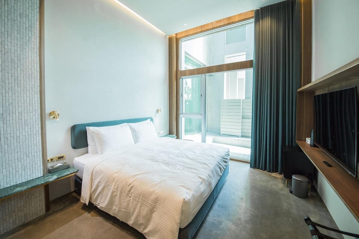 A modern hotel room with a large bed, a nightstand, a TV, and a sliding glass door to the outside.