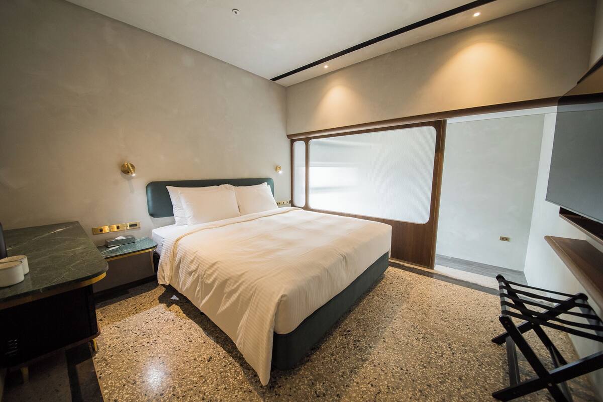 A modern hotel room with a large bed, a desk with a chair, and a window with blinds.