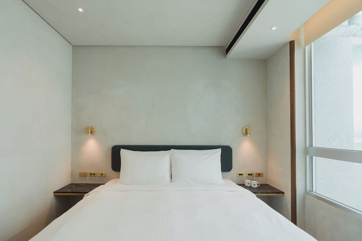 A modern hotel room with a large bed, two bedside tables, and a window providing natural light.