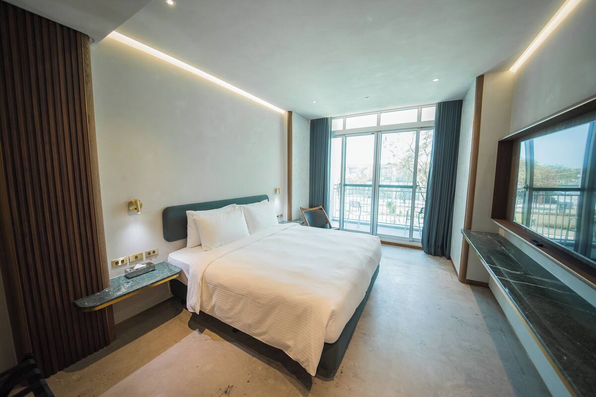 A modern hotel room with a large bed, a nightstand, a desk, and a view of the outdoors through large windows.
