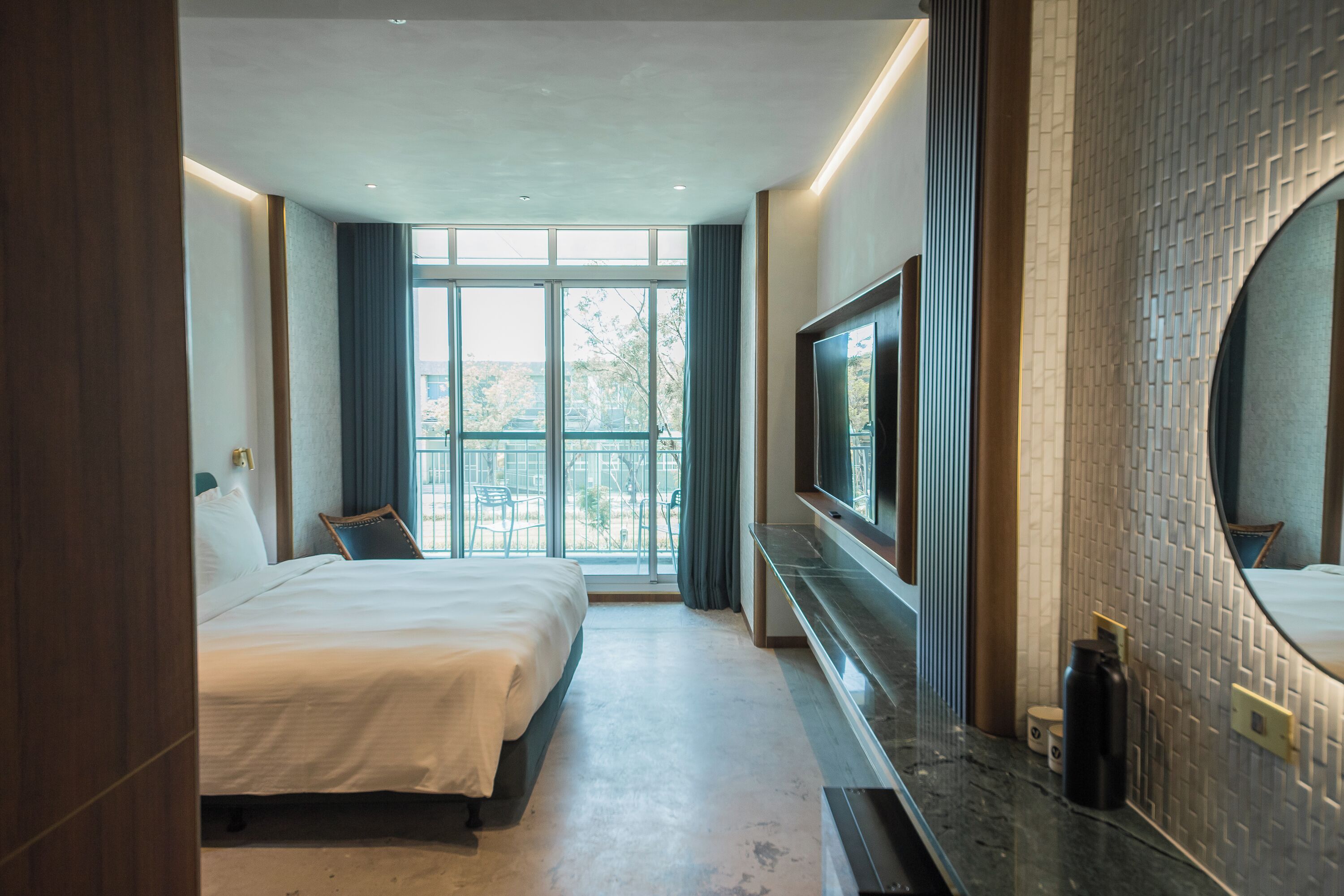 A hotel room with a large bed, a TV mounted on the wall, a bathroom with a mirror, and a view of the outdoors through the window.