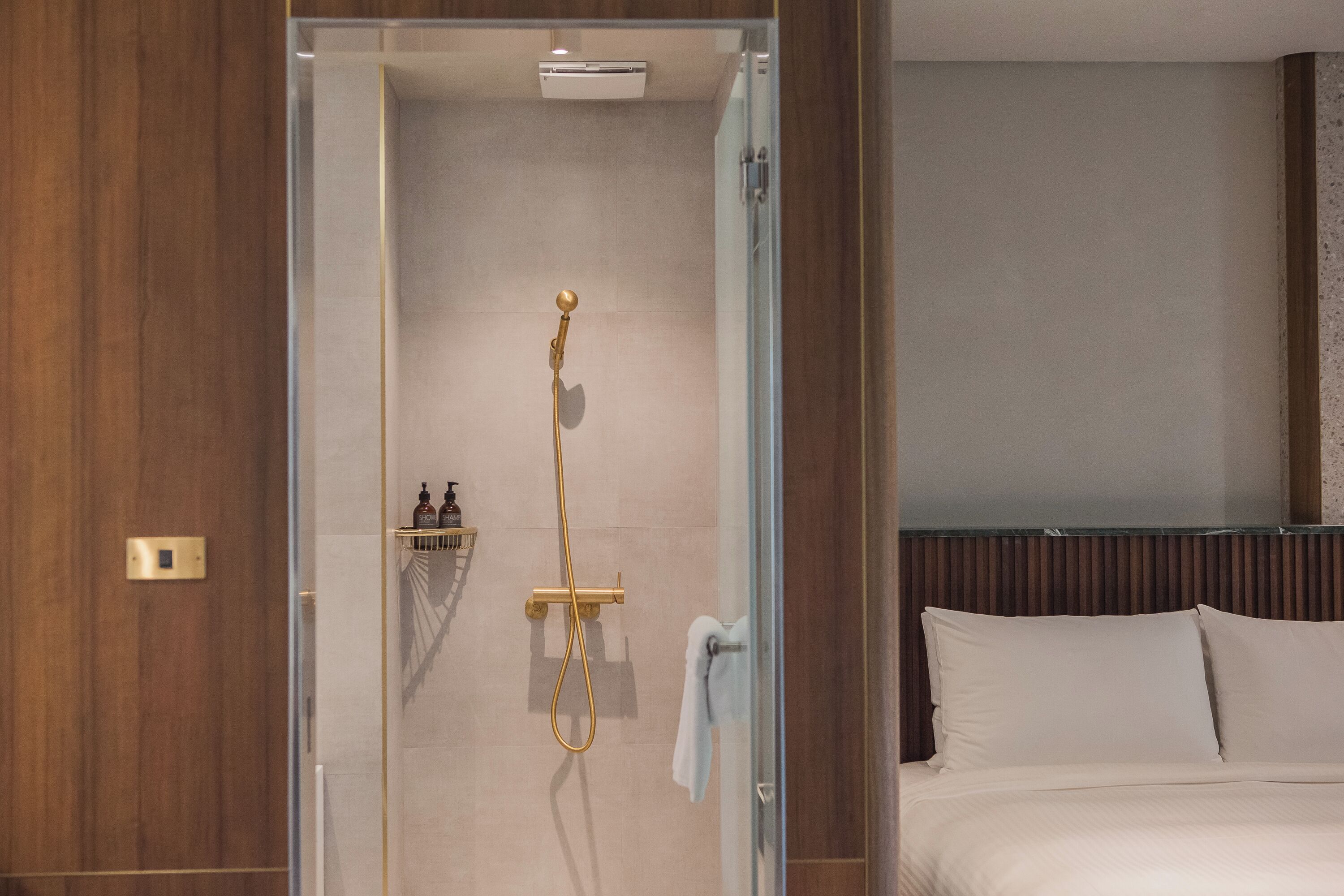 A hotel room with a glass shower enclosure, a bed with white linens, and a wooden headboard.