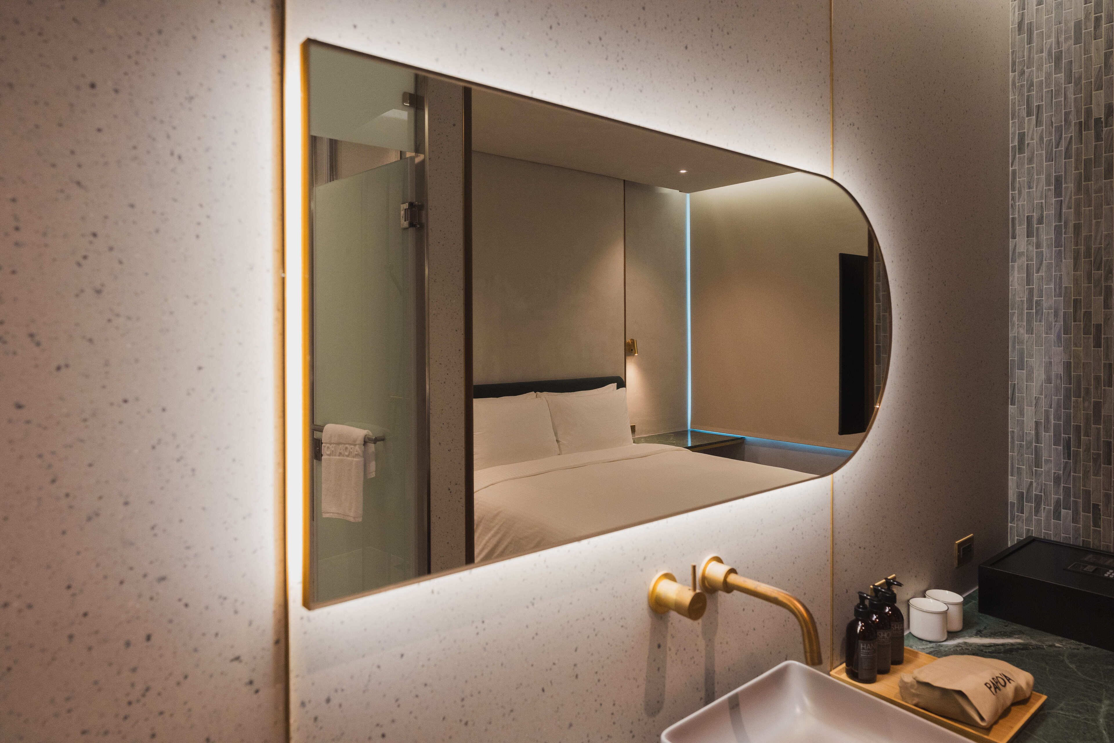 A modern hotel room with a large mirror, a bed with white linens, a bathroom with a sink and toiletries, and a tiled wall.