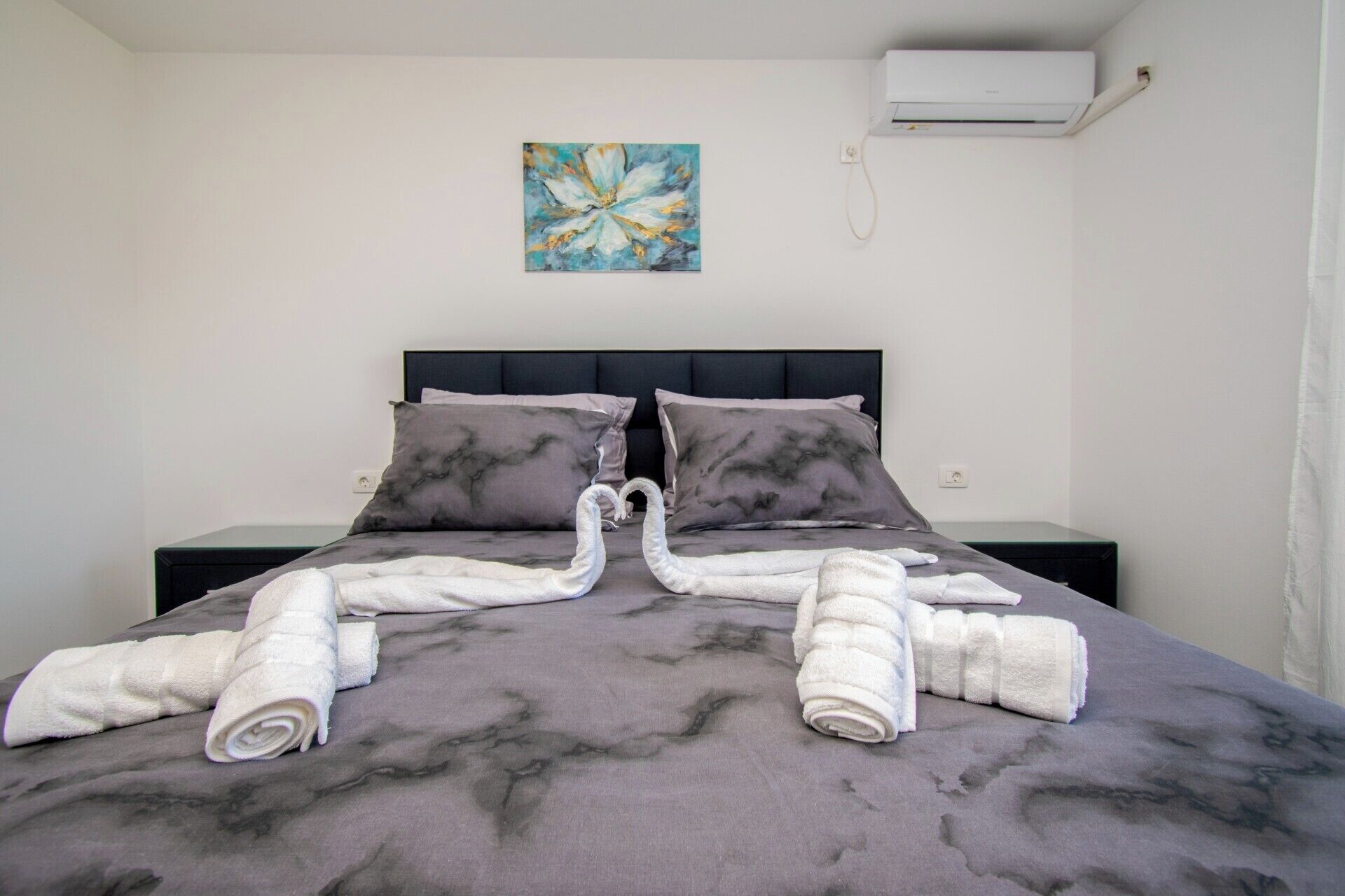 A neatly made bed with a marble-patterned bedspread, two pillows, and two rolled towels arranged in a heart shape.