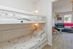 A bunk bed with a pillow and a bedside lamp, a sofa with a cushion, a small table, and a chair in a room with a window and artwork on the wall.