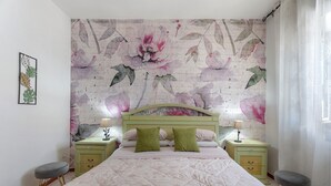 Ein Schlafzimmer mit einem Bett, zwei Nachttischen, einem Wandregal, einer Wand mit Blumenmuster-Tapete und einem Fenster mit Vorhängen.
