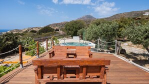 Una terraza de madera con piscina, bancos y vistas al mar y colinas.