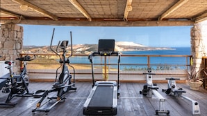 Ett utomhusgym med träningsredskap, ett trädäck och en vacker utsikt över havet och bergen.