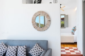 Une pièce avec un canapé gris, des coussins à motifs, un miroir rond et un lit posé sur un tapis à carreaux.