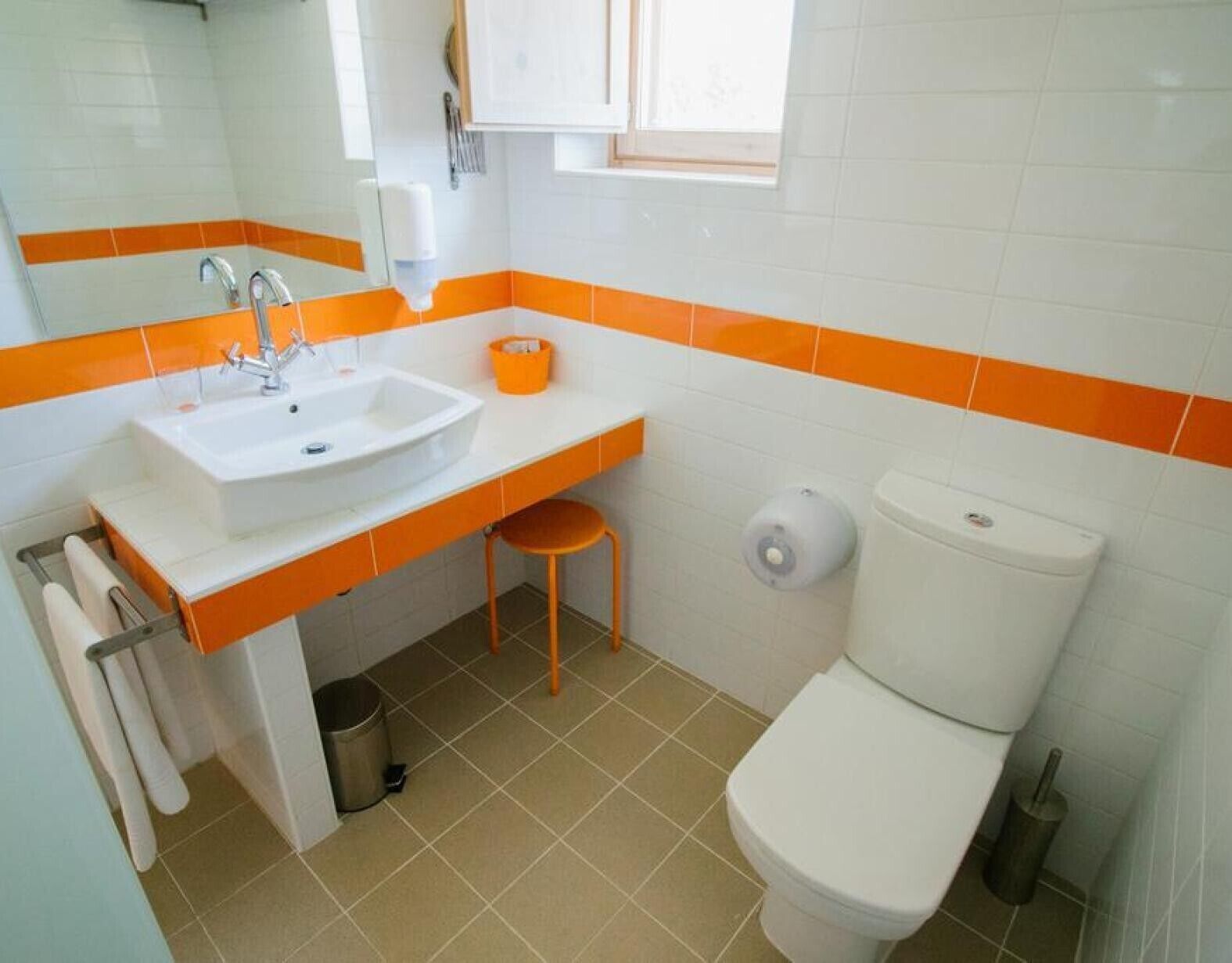 Casa de banho com lavatório branco, detalhes laranja, sanita e janela.