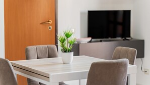 A modern dining area with a white table, four chairs, a potted plant, and a TV in the background.