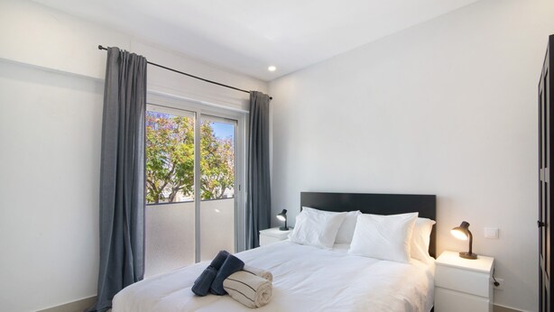Habitación de hotel con una cama grande, una mesita de noche con lámpara y vistas a árboles por la ventana.
