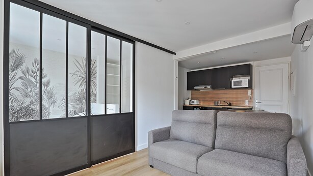 Una sala moderna con un sofá gris, un ventanal corredizo con vistas a un paisaje tropical, y una zona de cocina con armarios oscuros y un microondas.