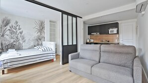 Una sala moderna con un sofá gris, una cama a rayas y una cocina con microondas y armarios de madera.