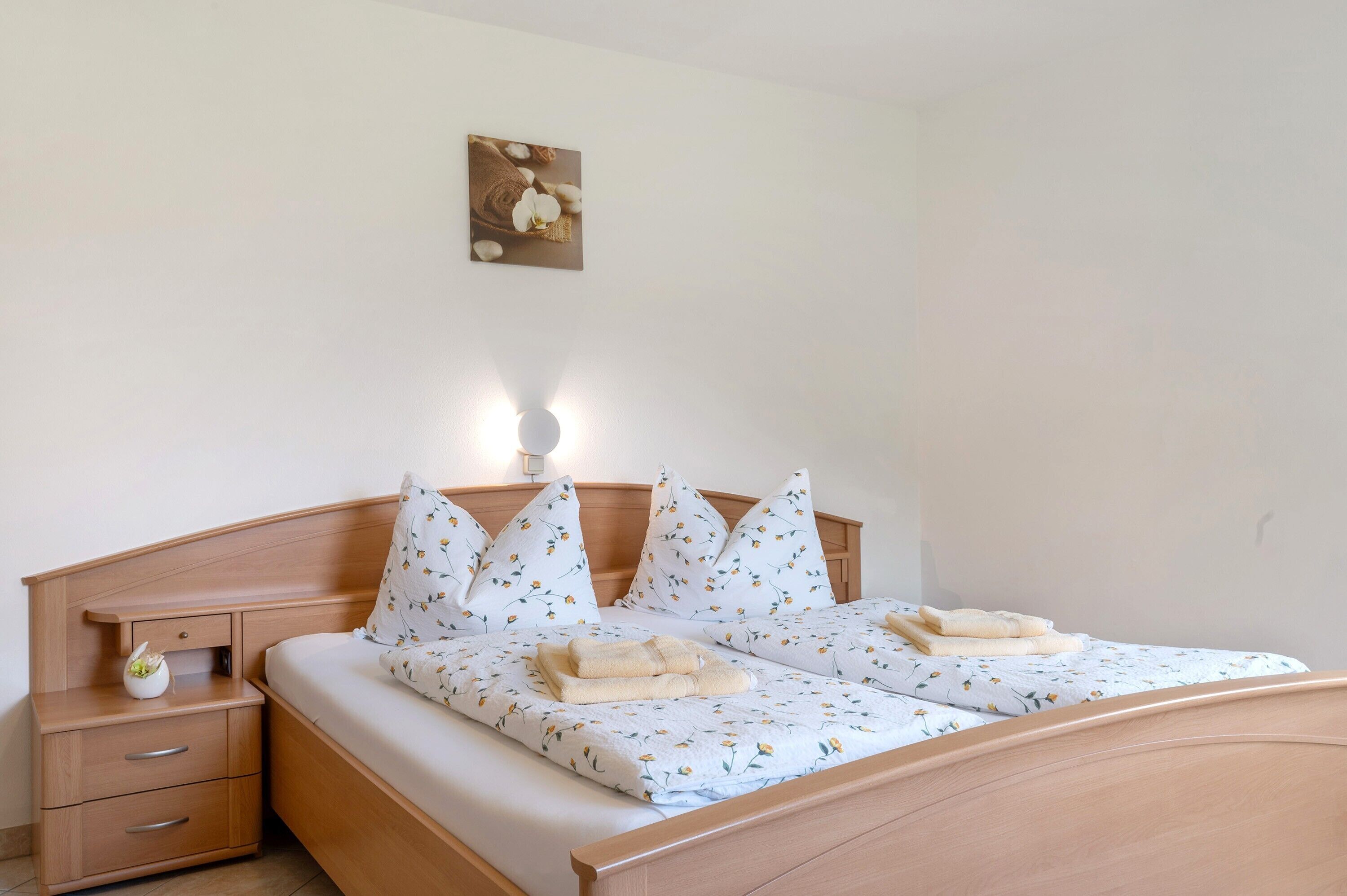 Een tweepersoonsbed met een bloemenprintbedspreider, twee kussens, een nachtkastje met een lamp en een aan de muur hangend schilderij.