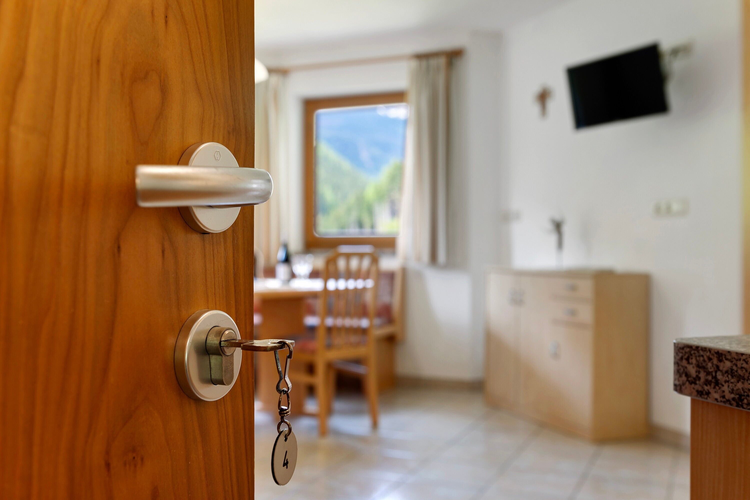 A wooden door with a keyhole, a dining area with a table and chairs, a window with a view of greenery, and a television mounted on the wall.
