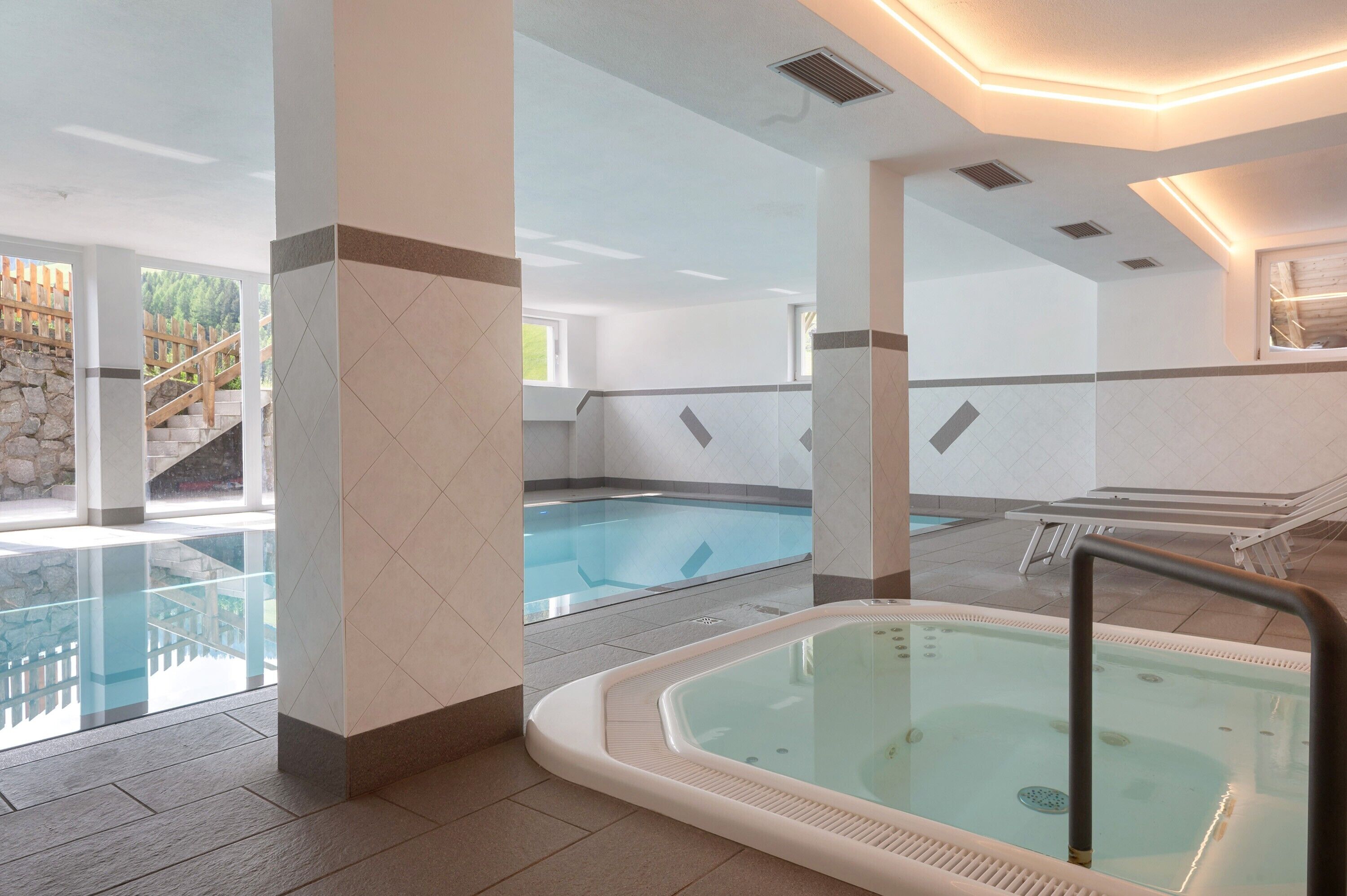 A hotel indoor pool area with a hot tub, lounge chairs, and a staircase leading outside.