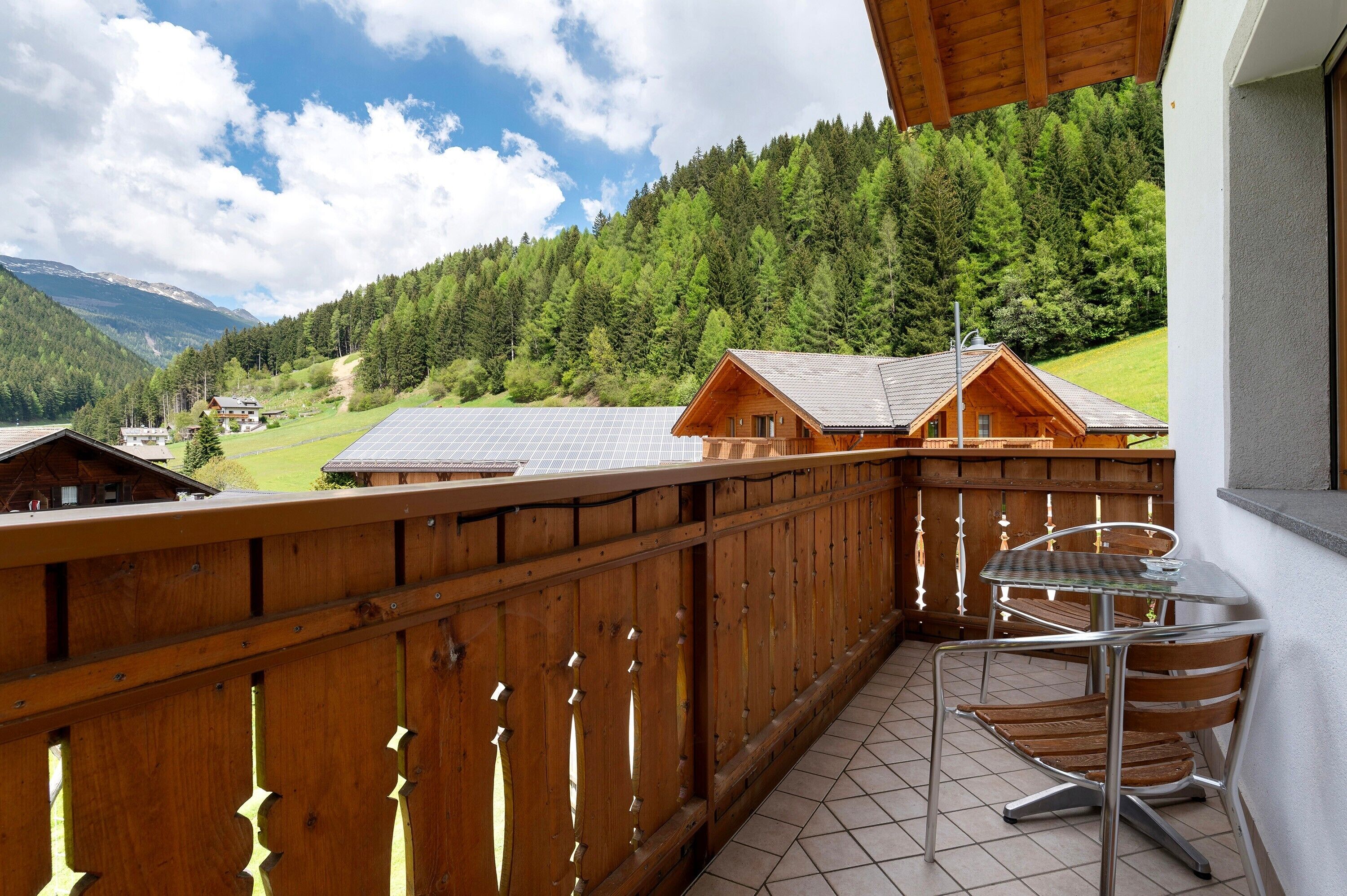 A wooden balcony with a table and chairs, overlooking a forested mountain area.