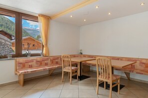 Ein Eckzimmer mit einer hölzernen Sitzbank, Tisch und Stühlen, einem Fenster mit Vorhang und Blick auf einen Berg.