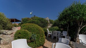 Un espace patio avec des chaises et des tables en plastique blanc, entouré de verdure et d’une formation rocheuse.