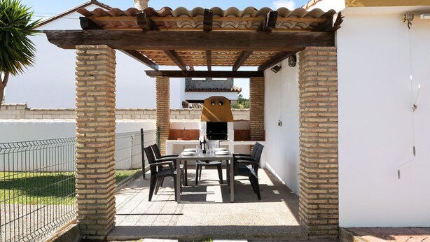 Une terrasse couverte avec un barbecue en briques, une table avec des chaises et un mur blanc.