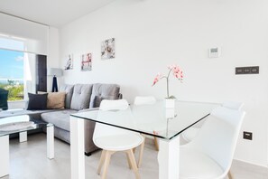 Un salon moderne avec une table à manger en verre, des chaises blanches, un canapé gris et une vue sur l’extérieur.