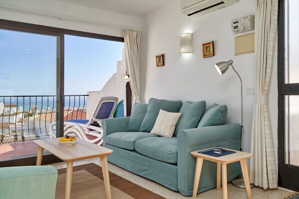 A balcony with a view of the sea, a teal sofa, a small table with a fruit bowl, and a lamp.