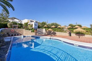 A clear blue pool with a stone wall and lifebuoys, surrounded by a stone pathway and thatched umbrellas.