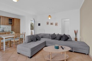 Ein modernes Wohnzimmer mit einem grauen Ecksofa, einem runden Couchtisch und einem Essbereich mit weißem Tisch und Stühlen.