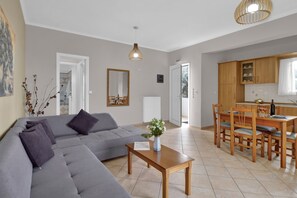 A modern living room with a grey sofa, a wooden dining table, and a kitchen area with wooden cabinets and a window.